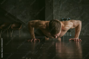 Young sexy male bodybuilder athlete with a bare torso. portrait of a studio of a light-skinned strong man engaged in push-ups, on the floor, straining powerful hands